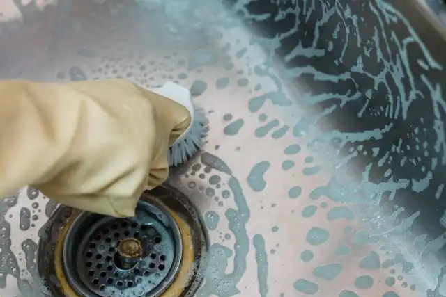 Mão com luva amarela esfrega uma pia de inox com escova e sabão