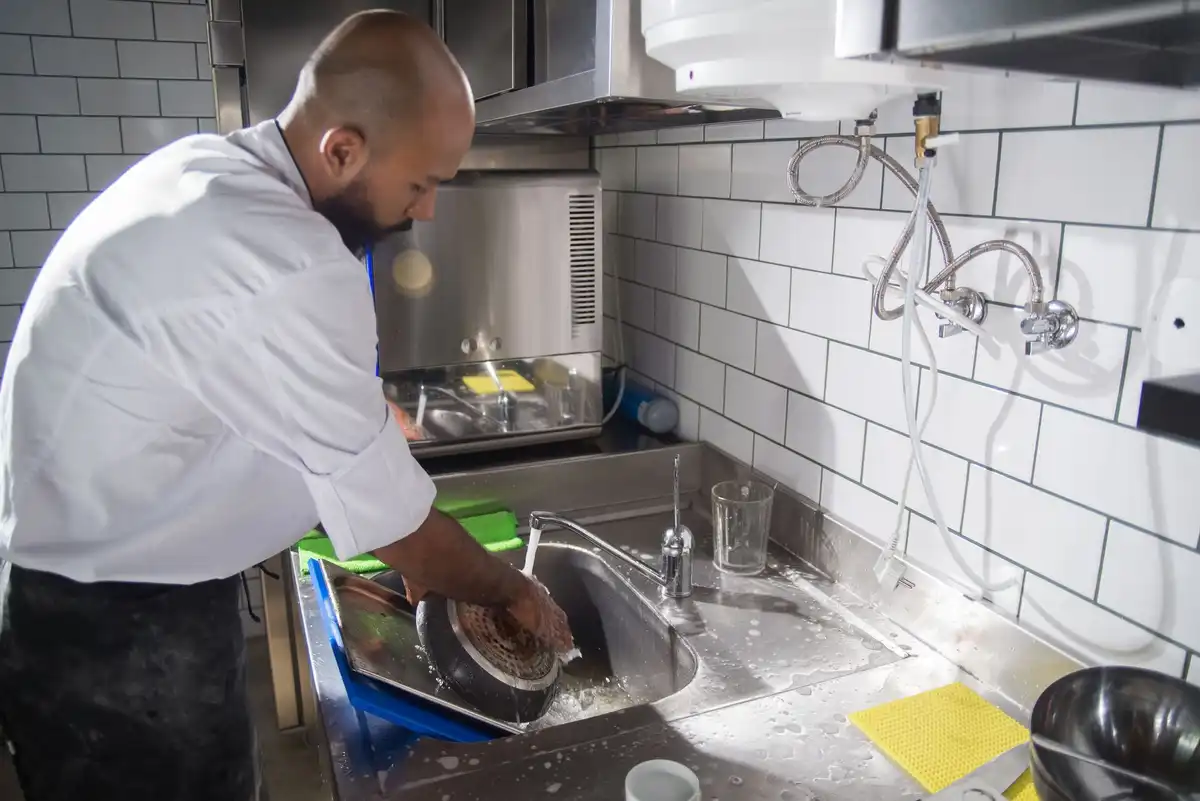 Homem lava uma frigideira na pia de inox de uma cozinha comercial